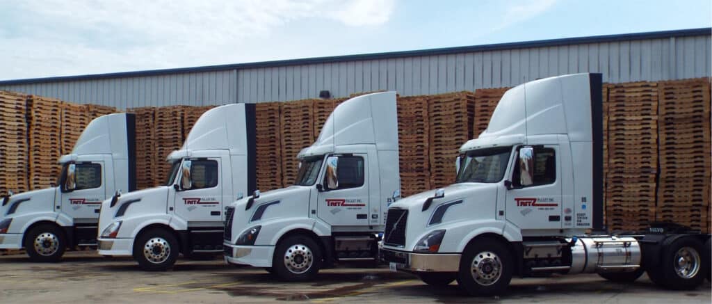 Four white semi-trucks parked in front of wooden pallets