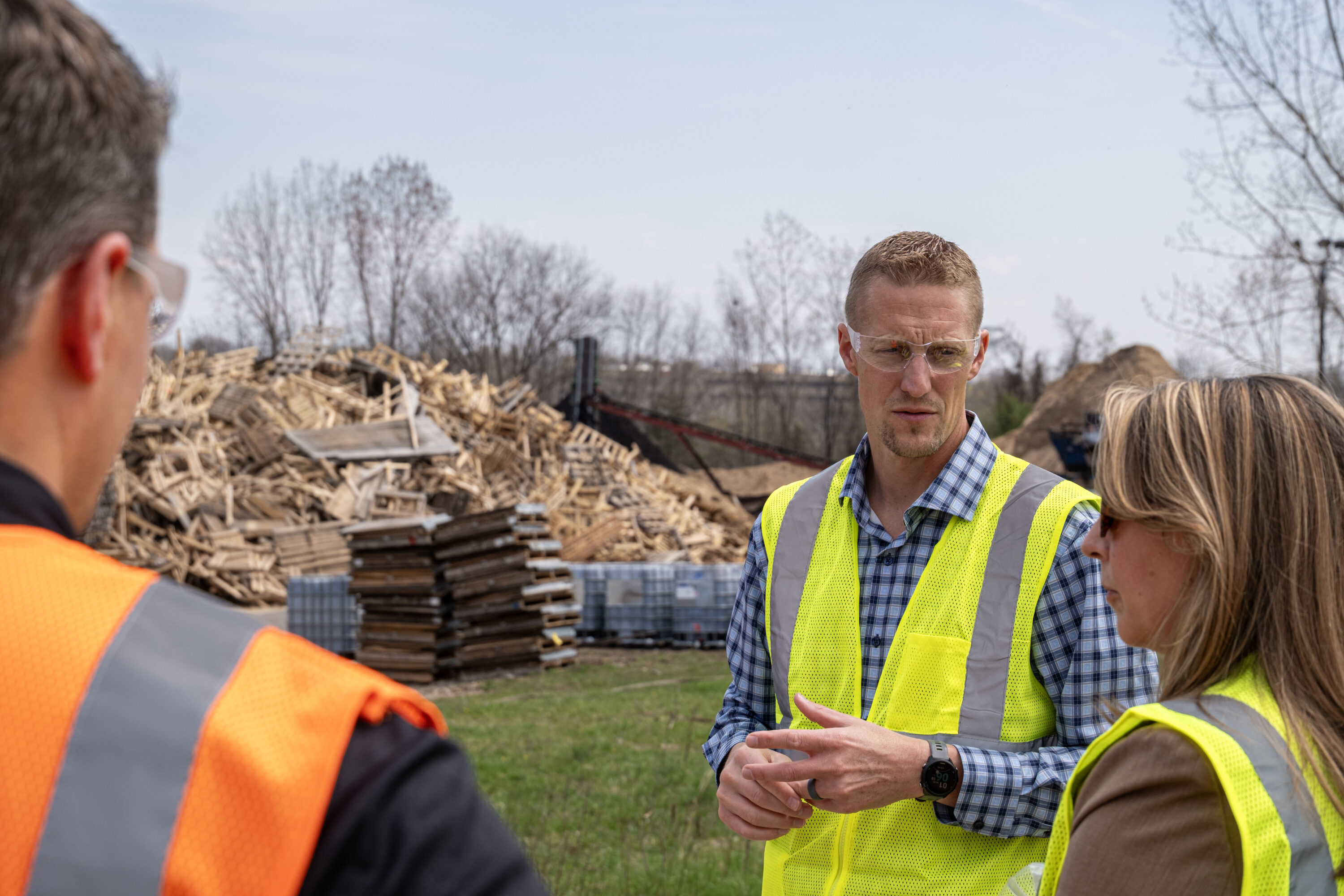 A man wearing a high visibility vest and safety glasses stares and listens intently to woman in front of large scrap pile of used wooden pallets.