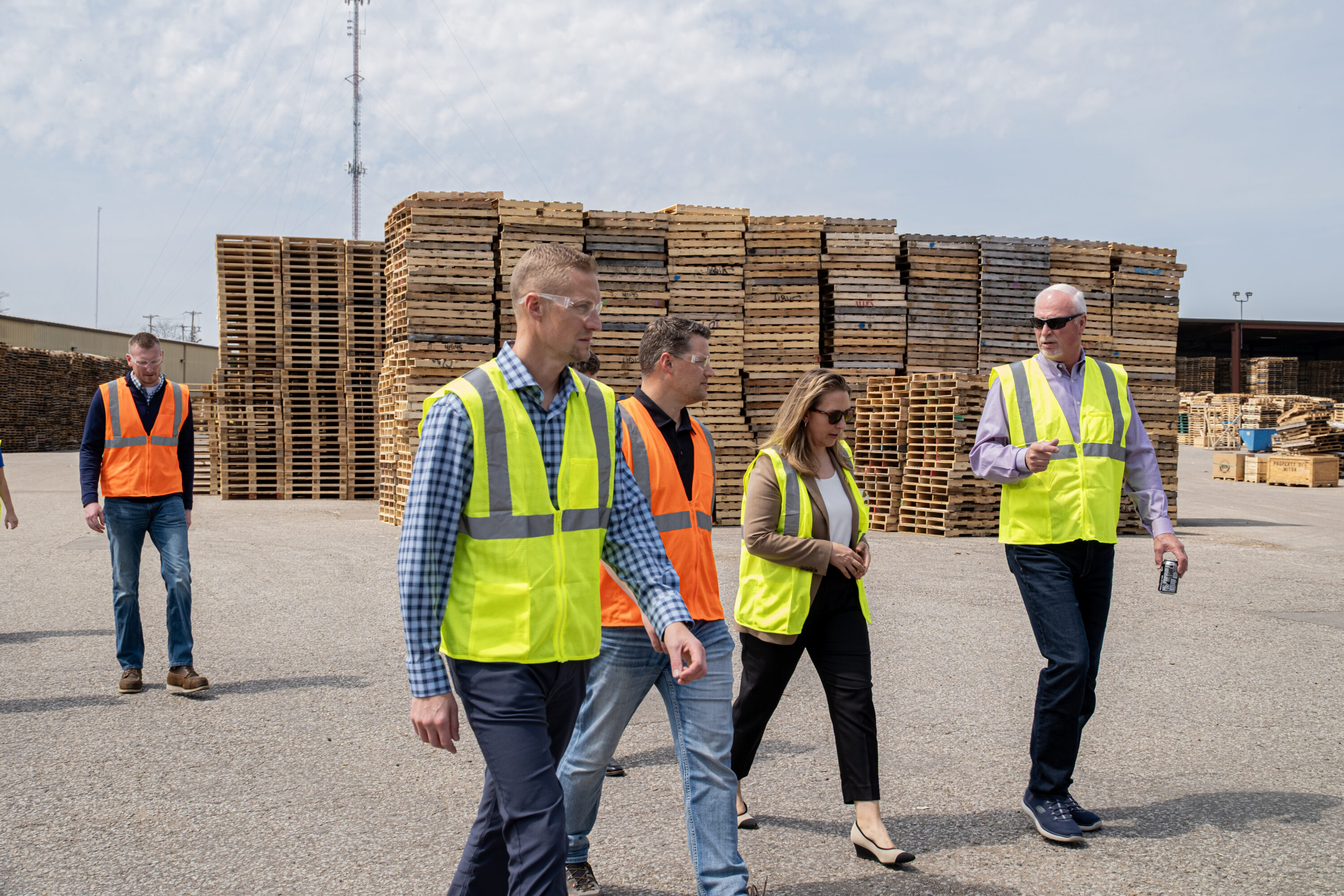 A group of men and women wearing high visibility vests walking in pallet yard with a large stack of wooden pallets behind them