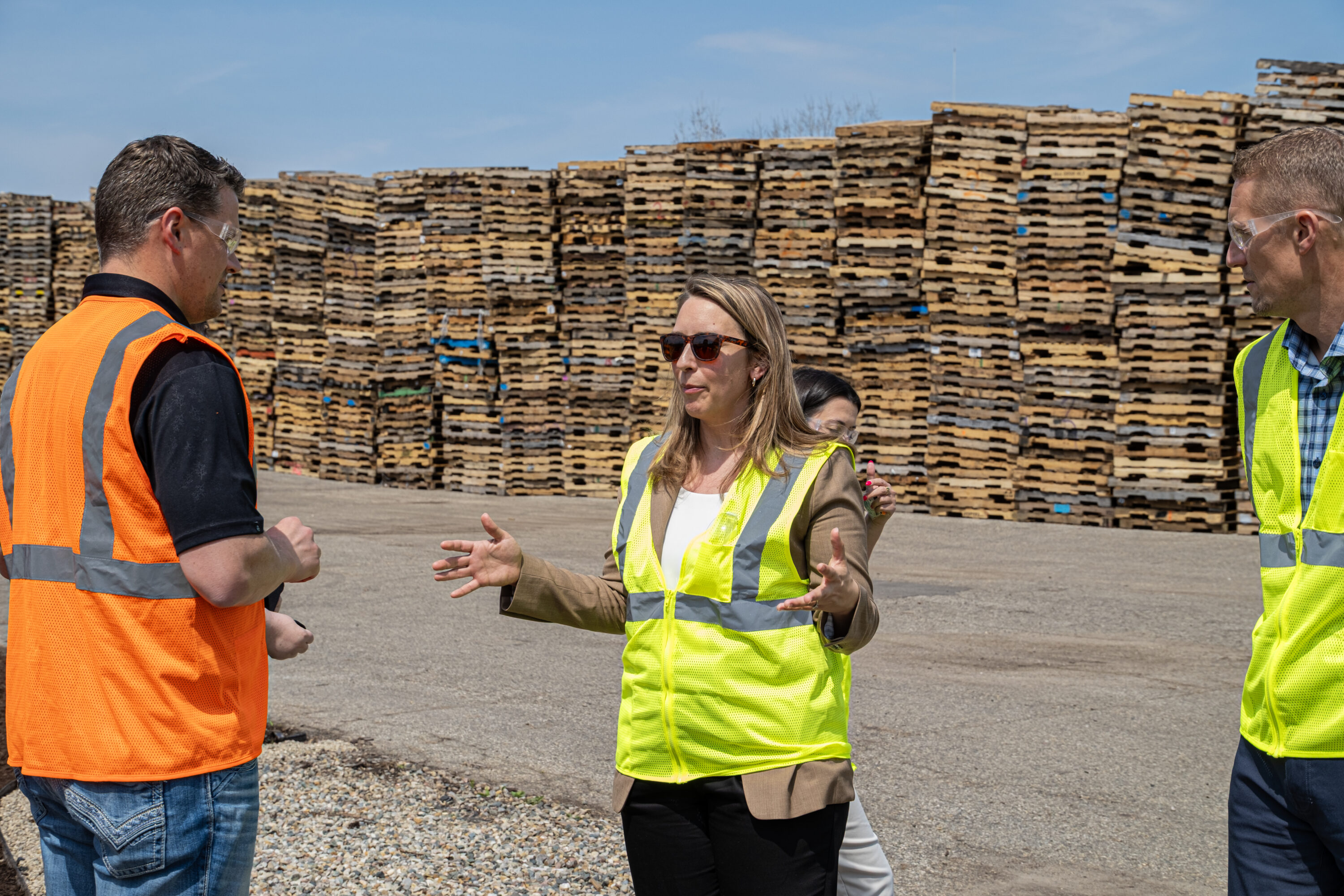 A group of men and women wearing high visibility vests talking in pallet yard with a large stack of wooden pallets behind them.