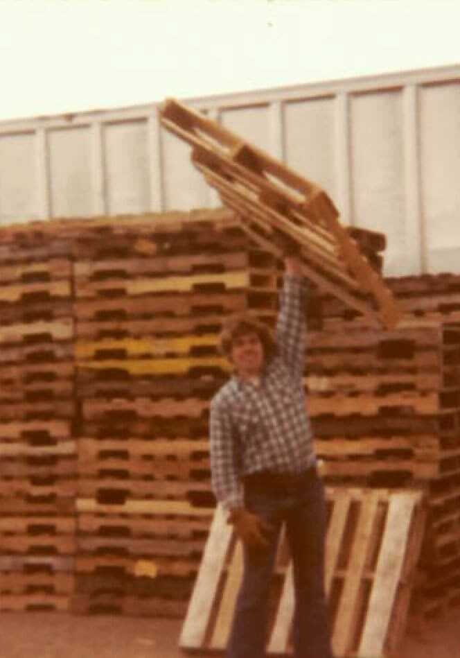Strong man in old photo holding a pallet above his head with one arm.